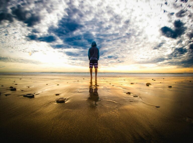 A lone figure stands on a tranquil beach during a vivid sunset, reflecting on the wet sand.