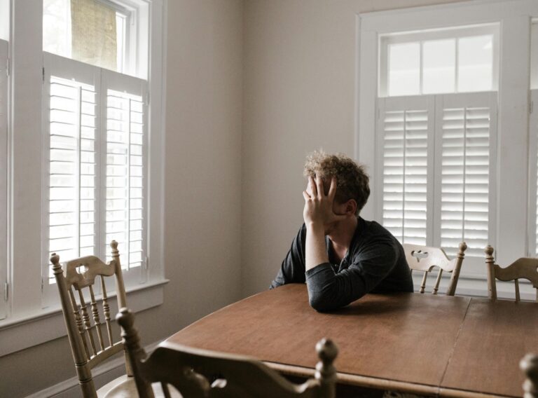 A man sits alone at a table in a bright room, displaying deep contemplation.