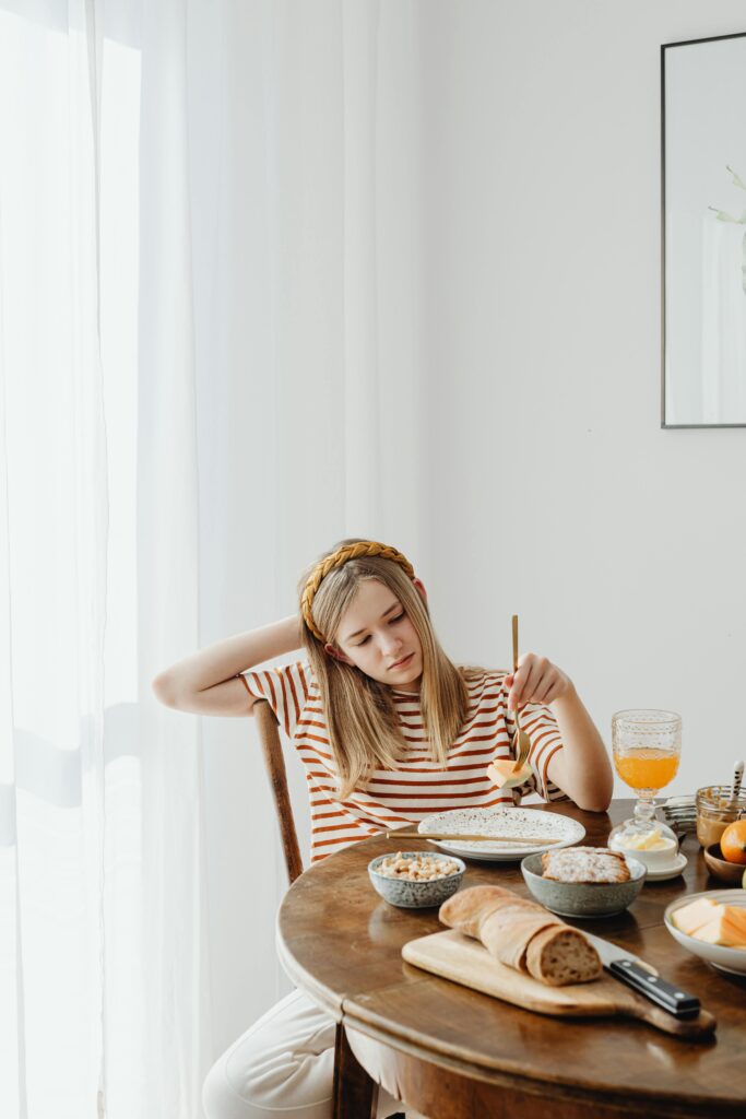 A teenage girl sits dissatisfied at breakfast with various food items on a wooden table.