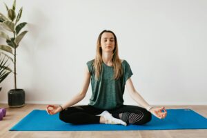 Serene blond lady in sportswear sitting with crossed legs and closed eyes while practicing yoga at home near potted plants