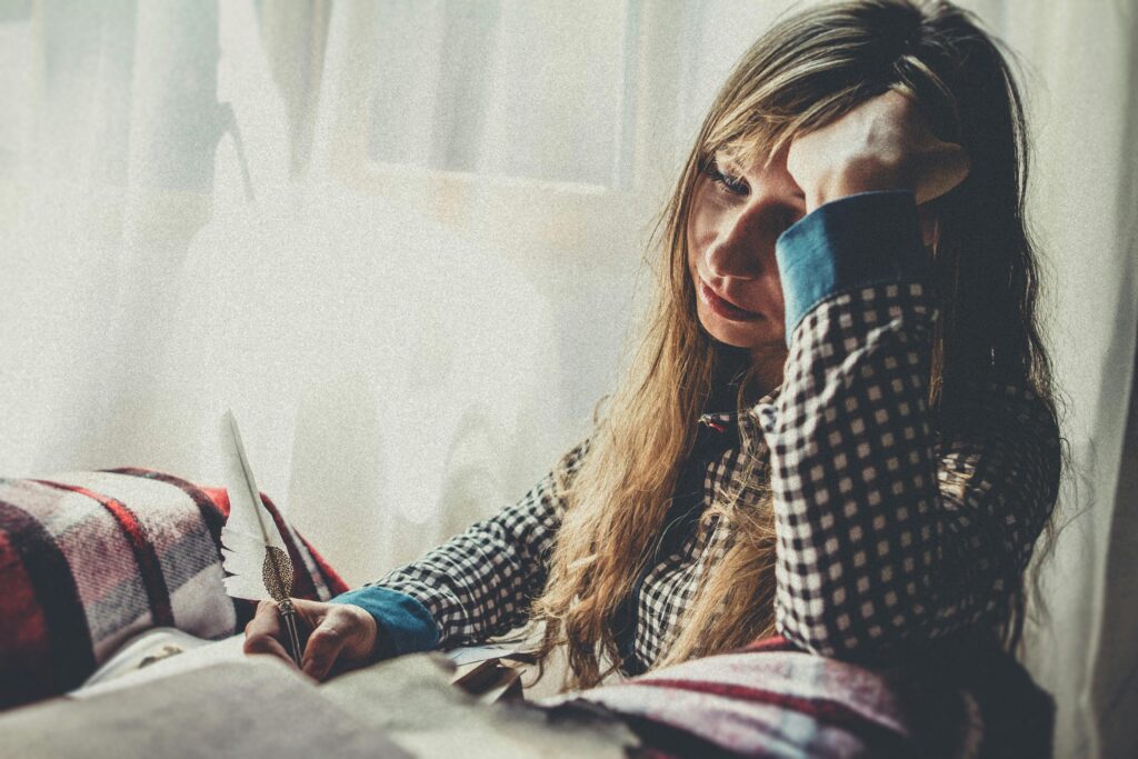 A woman with a quill, deep in thought by a sunlit window, in a plaid shirt.
