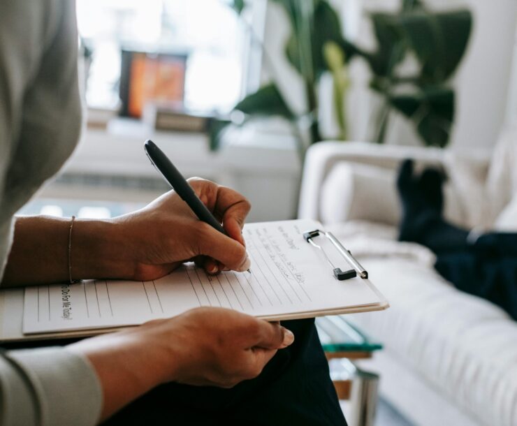 Unrecognizable ethnic female therapist taking notes on clipboard while filling out form during psychological appointment with anonymous client lying on blurred background
