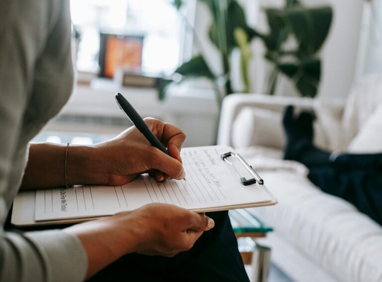pexels-photo-5699456-5699456 Unrecognizable ethnic female therapist taking notes on clipboard while filling out form during psychological appointment with anonymous client lying on blurred background